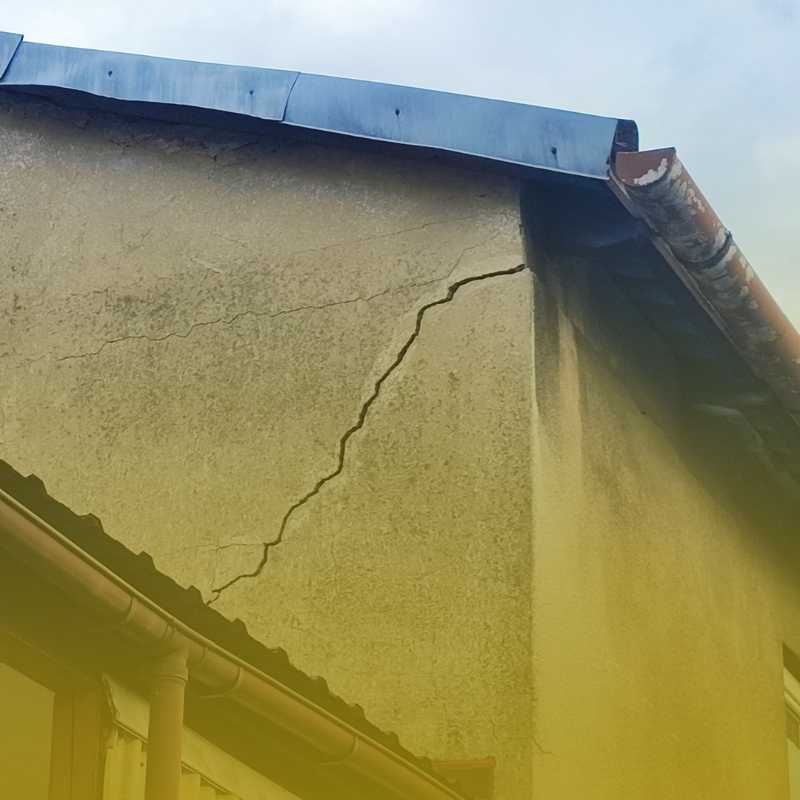 Zbliżenie narożnika dachu i rynny; głębokie pęknięcie biegnące ukośnie po elewacji, żółty gradient na dole kadru.