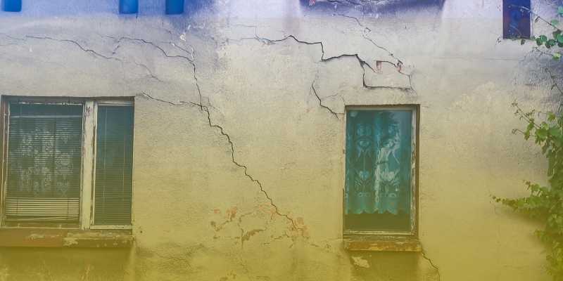 Fasada starego domu z dwoma oknami; liczne pęknięcia i odspojenia tynku, żółty gradient przy dolnej krawędzi.