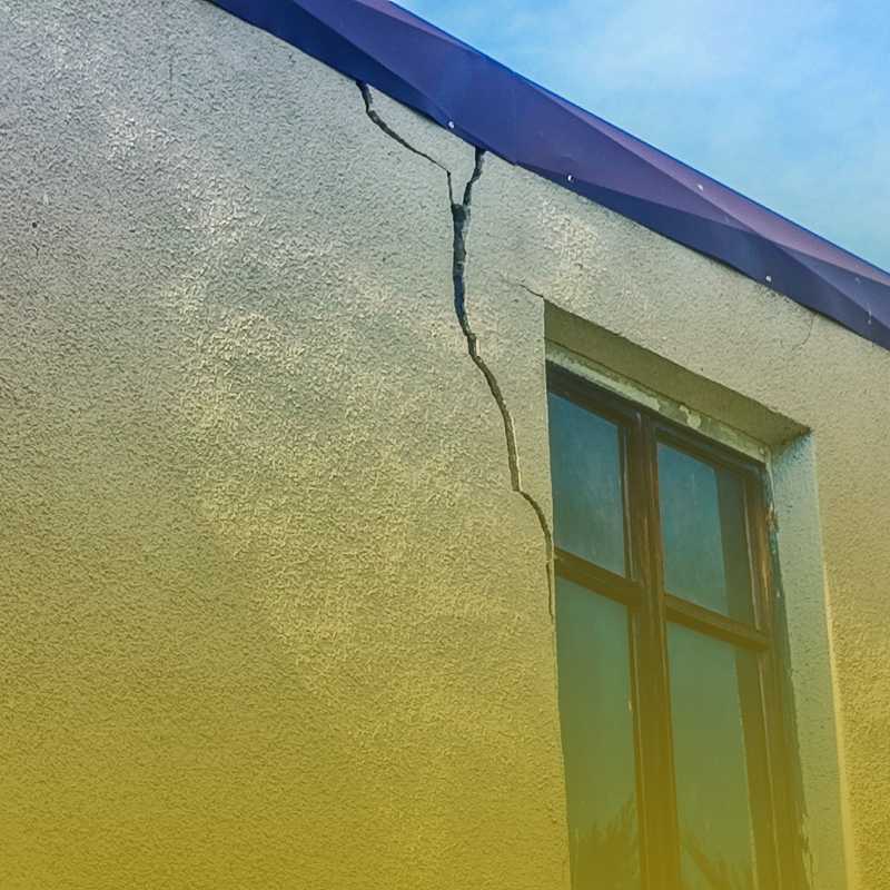 Narożnik budynku z oknem; pionowe pęknięcie od górnej krawędzi ściany w dół, lekki żółty gradient.